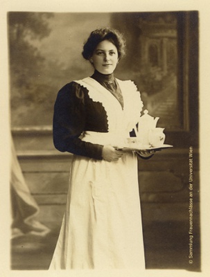 Martha Teichmann in schwarzer Bluse und weißer Schürze mit einem Tablett mit Teegeschirr in der Hand